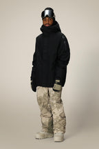 A person stands against a plain background wearing a black hooded jacket, 686 Mens GORE-TEX Core Shell Pant in tan and gray camo, light gray gloves, beige snow boots, and black ski goggles on their forehead.