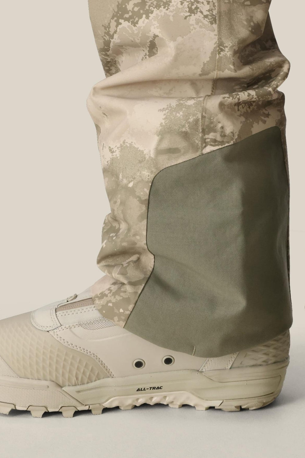 Close-up of a person wearing 686 Mens GORE-TEX Core Shell Pant in beige and green, with a reinforced patch over a beige rugged hiking boot, set against a light background.