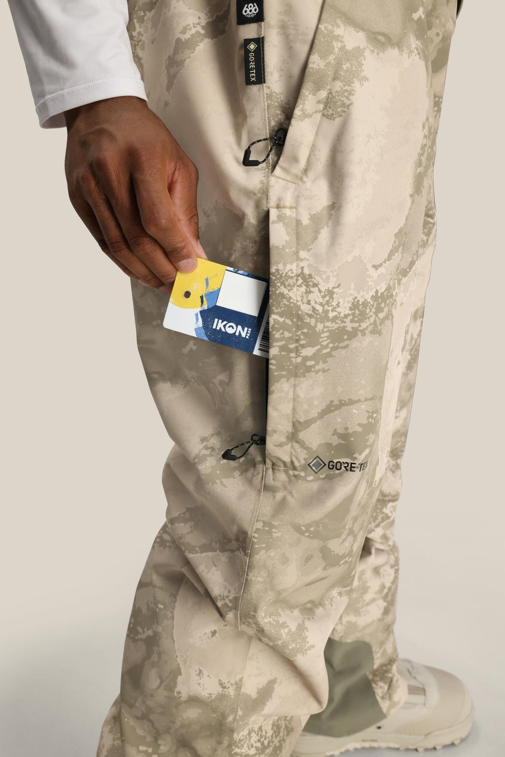 A person slips a blue and white IKON card into the side pocket of beige camo 686 Mens GORE-TEX Core Shell Pant. Only their lower body and hand are seen.