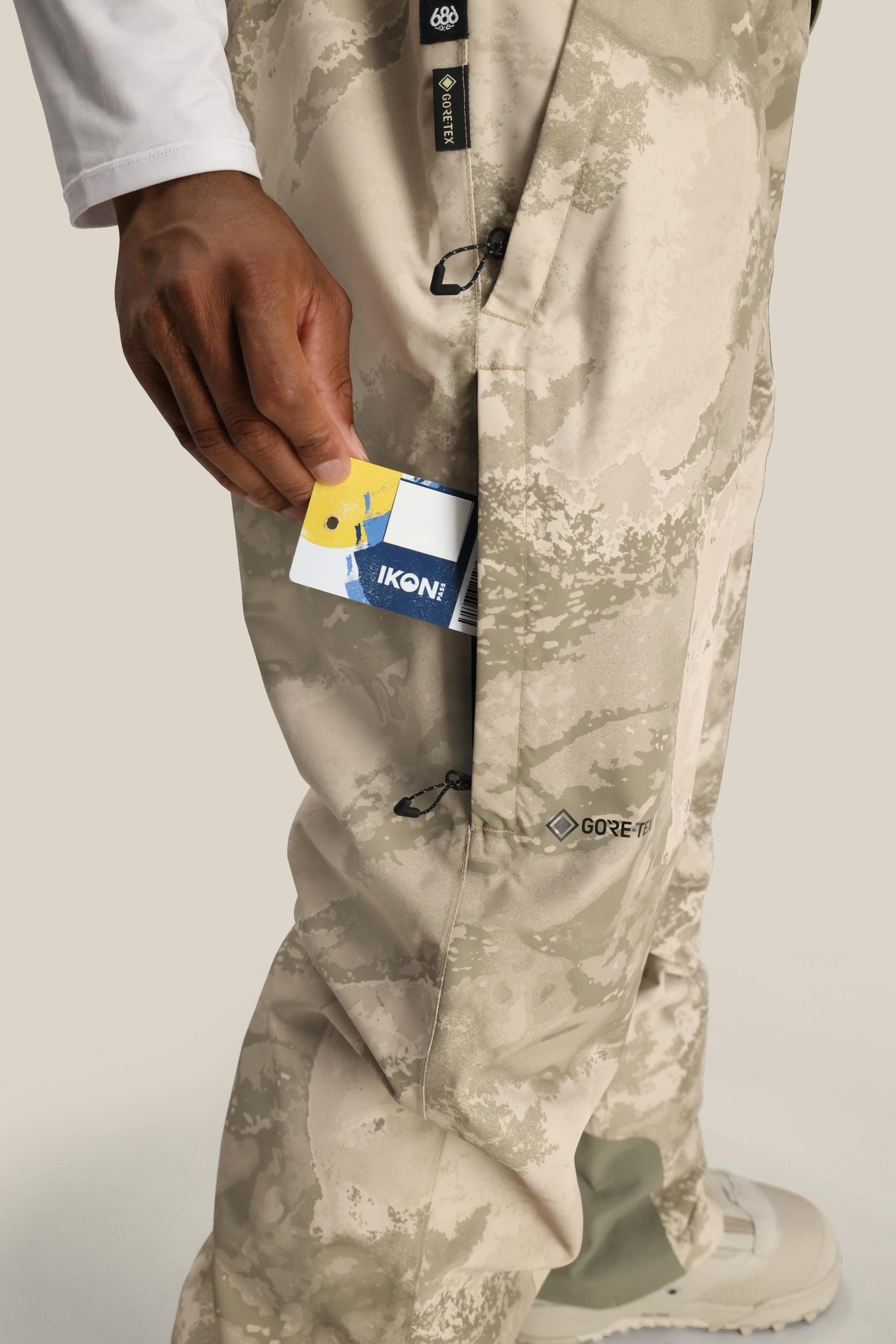 A person slips a blue and white IKON card into the side pocket of beige camo 686 Mens GORE-TEX Core Shell Pant. Only their lower body and hand are seen.