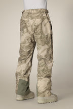 A person wearing 686 Mens GORE-TEX Core Shell Pant in light tan and brown, with pockets and reinforced green fabric on the lower legs, a white shirt tucked in, and beige boots, shown from the back.