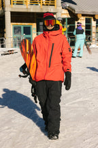 A snowboarder in a bright orange jacket, 686 Men's GORE-TEX Dispatch™ Shell Pant by 686, gloves, and goggles holds a snowboard while walking on snow near a ski lodge. Other people and snowboarding gear are visible in the background.