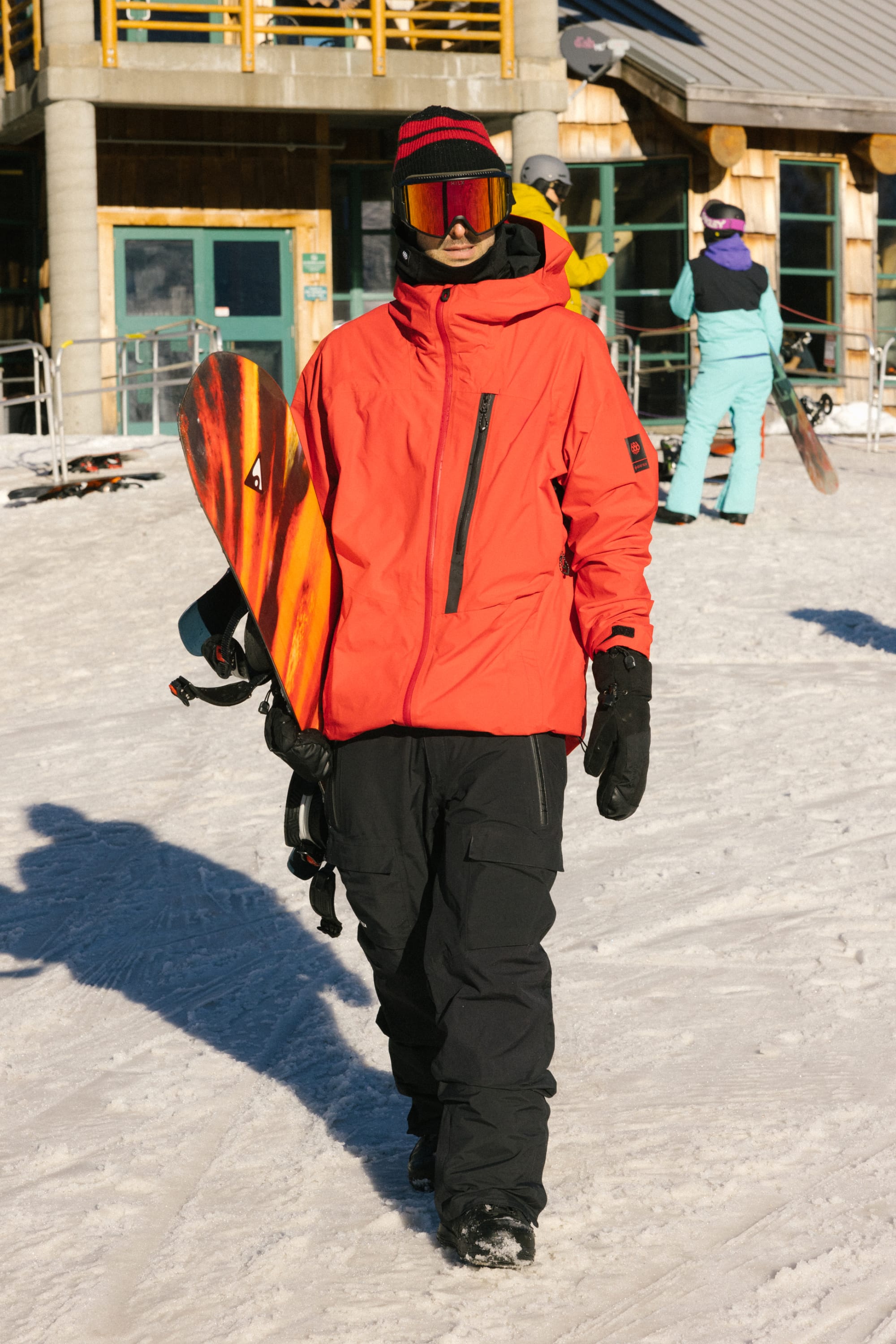 A snowboarder in a bright orange jacket, 686 Men's GORE-TEX Dispatch™ Shell Pant by 686, gloves, and goggles holds a snowboard while walking on snow near a ski lodge. Other people and snowboarding gear are visible in the background.