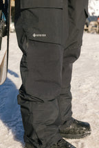 A close-up of a person wearing 686 Men's GORE-TEX Dispatch™ Shell Pant with black boots, standing on snow, and part of a snowboard is visible on the left.