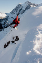 A snowboarder in a red jacket and 686 Men's GORE-TEX Dispatch™ Shell Pant jumps off a snowy slope, surrounded by snow-covered mountains under a clear sky.