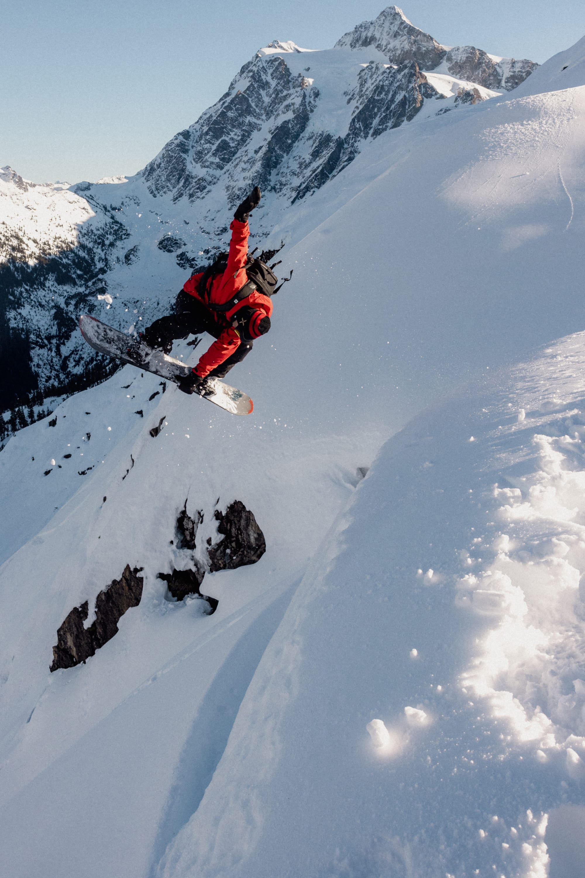 A snowboarder in a red jacket and 686 Men's GORE-TEX Dispatch™ Shell Pant jumps off a snowy slope, surrounded by snow-covered mountains under a clear sky.