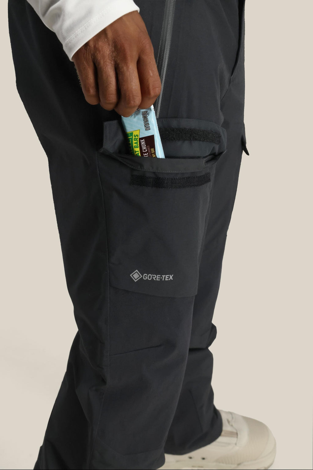 A person wearing 686 Men's GORE-TEX Dispatch™ Shell Pant in dark color puts a granola bar into a zippered side pocket. Only their lower body and hand are visible; they also wear white shoes and a white long-sleeve shirt.