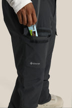 A person wearing 686 Men's GORE-TEX Dispatch™ Shell Pant in dark color puts a granola bar into a zippered side pocket. Only their lower body and hand are visible; they also wear white shoes and a white long-sleeve shirt.