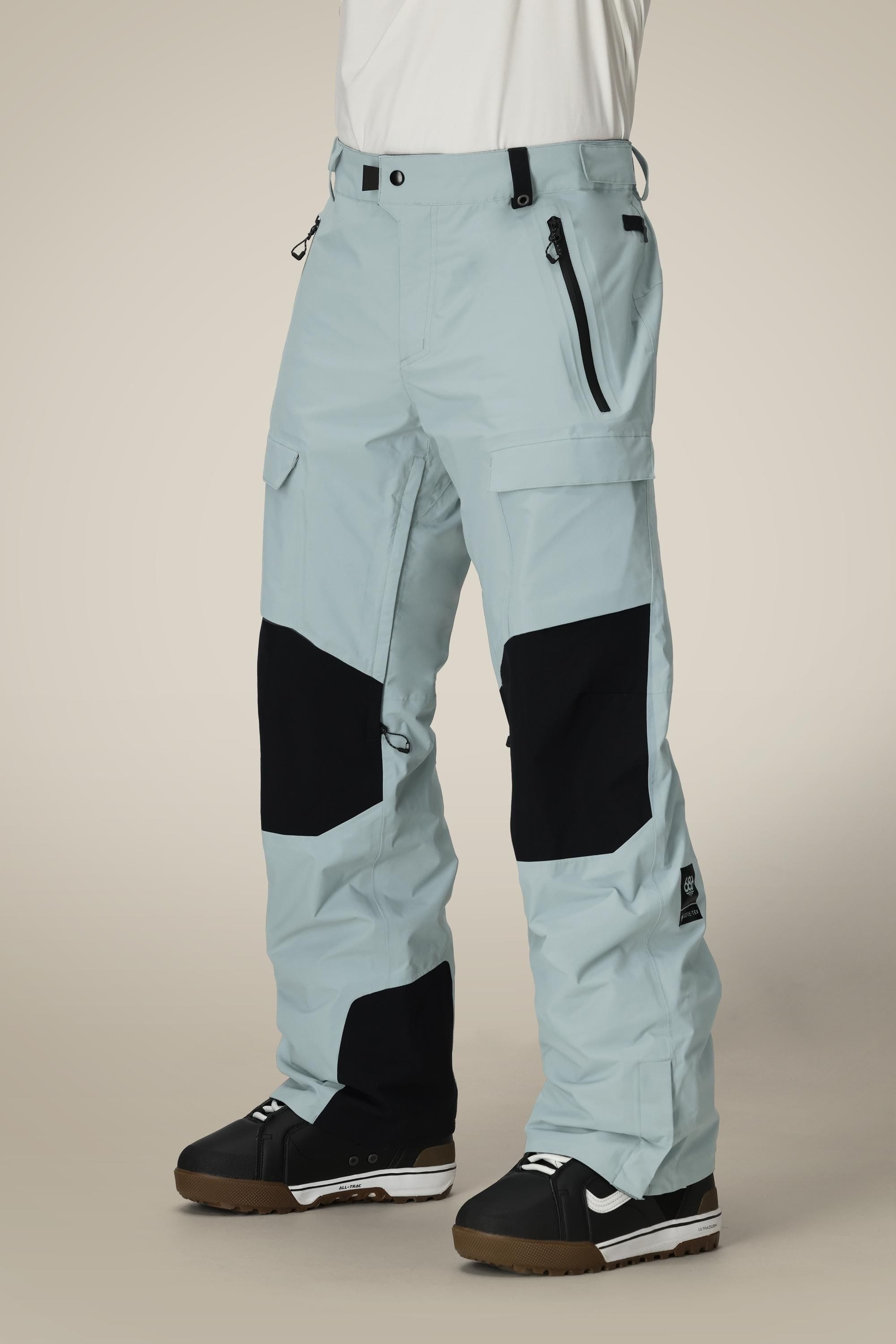 A person wearing 686 Men's GORE-TEX Dispatch™ Shell Pant in light blue with black knee patches and black-and-white winter boots, standing against a plain beige background. Only the lower half of their body is shown.