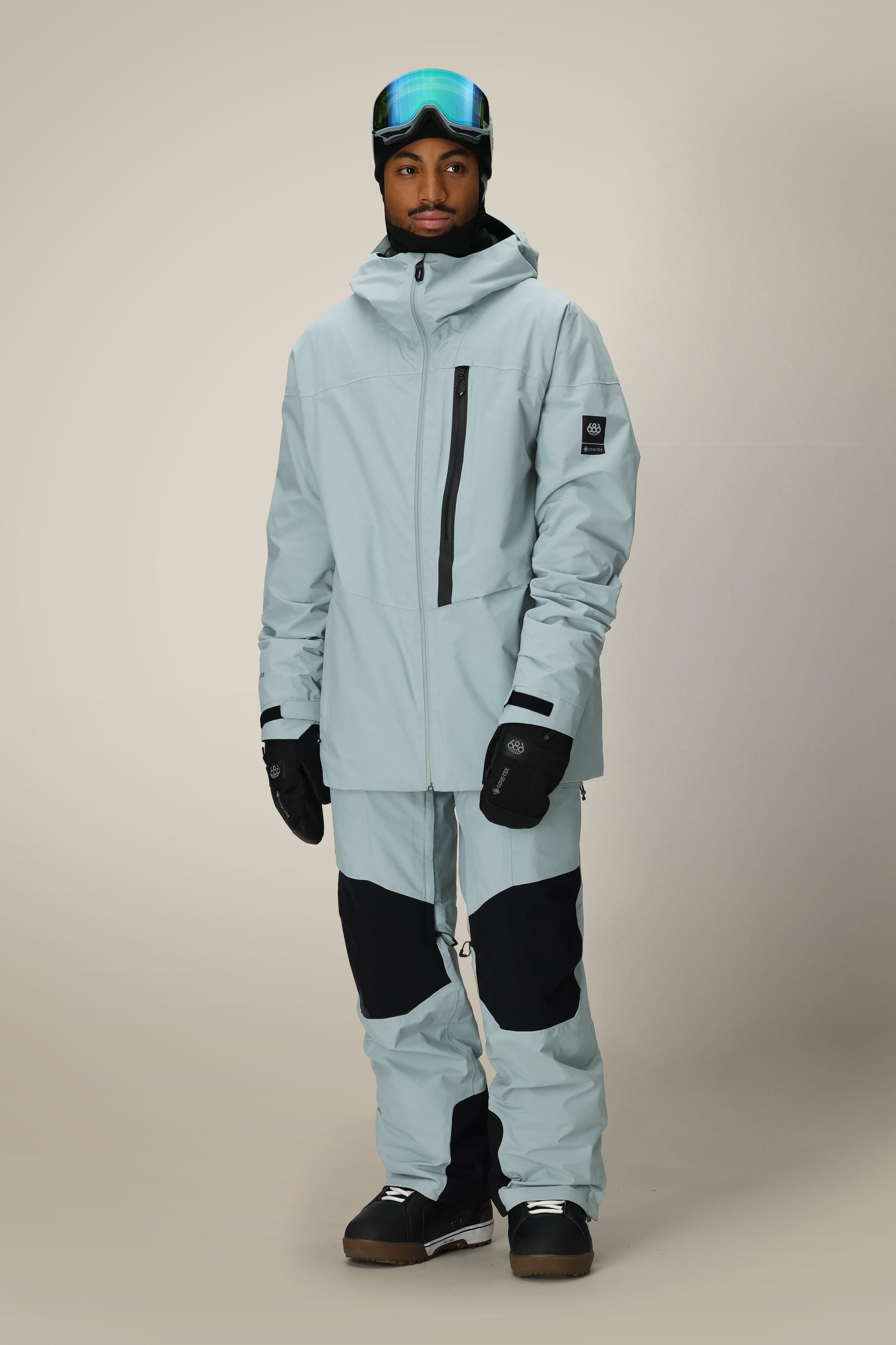 A person in a light blue ski jacket and 686 Men's GORE-TEX Dispatch™ Shell Pant stands against a plain beige background, wearing black gloves, dark boots, a black beanie, and ski goggles.