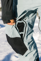 A person in 686 Men's GORE-TEX Dispatch™ Shell Pant stands outside in the snow, opening a zippered mesh thigh vent.