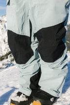 A close-up of a person wearing 686 Men's GORE-TEX Dispatch™ Shell Pant in light blue with black knee patches and black snow boots, standing on snow against a snowy mountain backdrop.