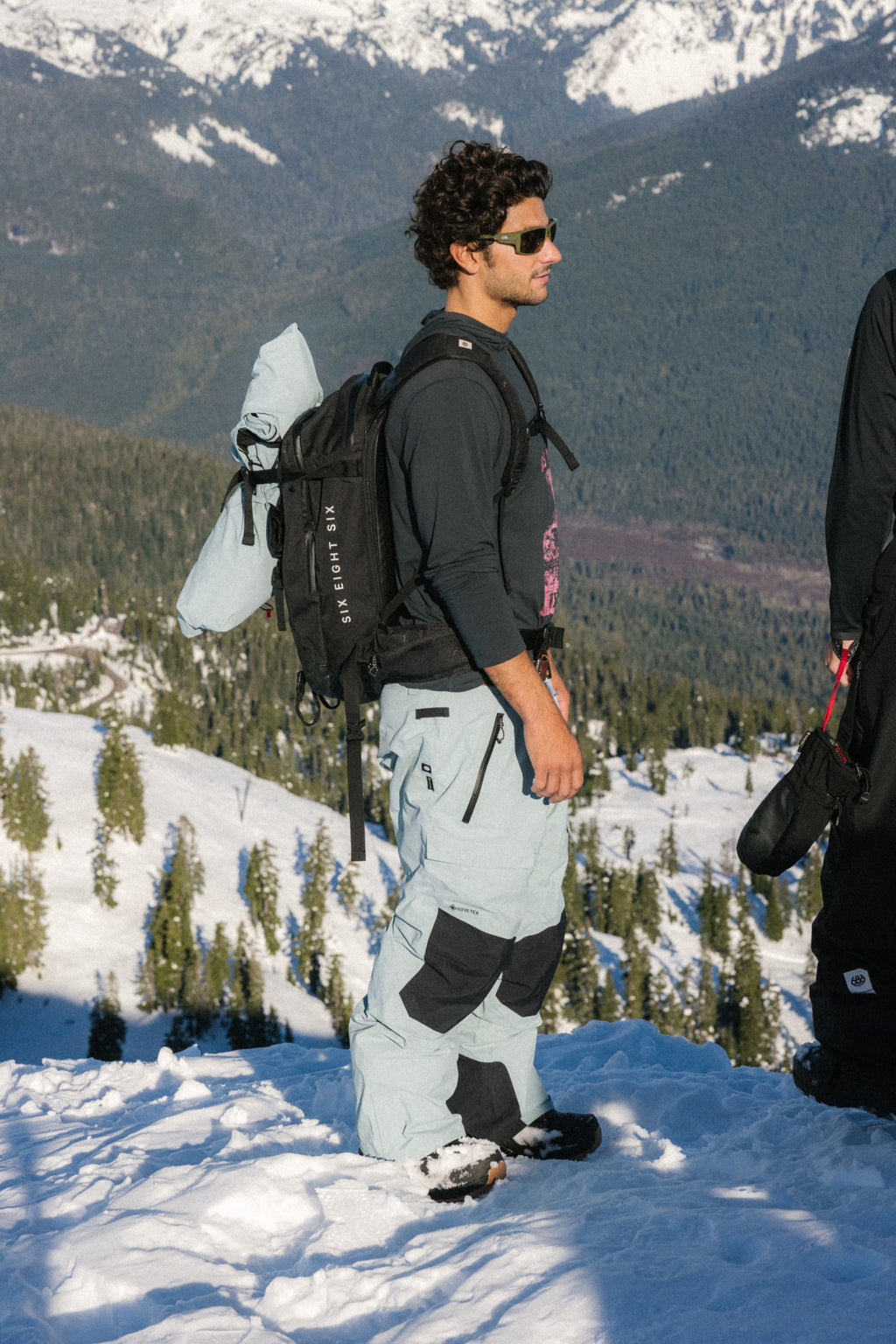 A man stands on a snowy mountain slope, wearing sunglasses, a black shirt, and the 686 Men's GORE-TEX Dispatch™ Shell Pant by 686. With a backpack and snowboard, he gazes ahead as pine trees and snow-capped peaks create a stunning backdrop.