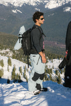 A man stands on a snowy mountain slope, wearing sunglasses, a black shirt, and the 686 Men's GORE-TEX Dispatch™ Shell Pant by 686. With a backpack and snowboard, he gazes ahead as pine trees and snow-capped peaks create a stunning backdrop.