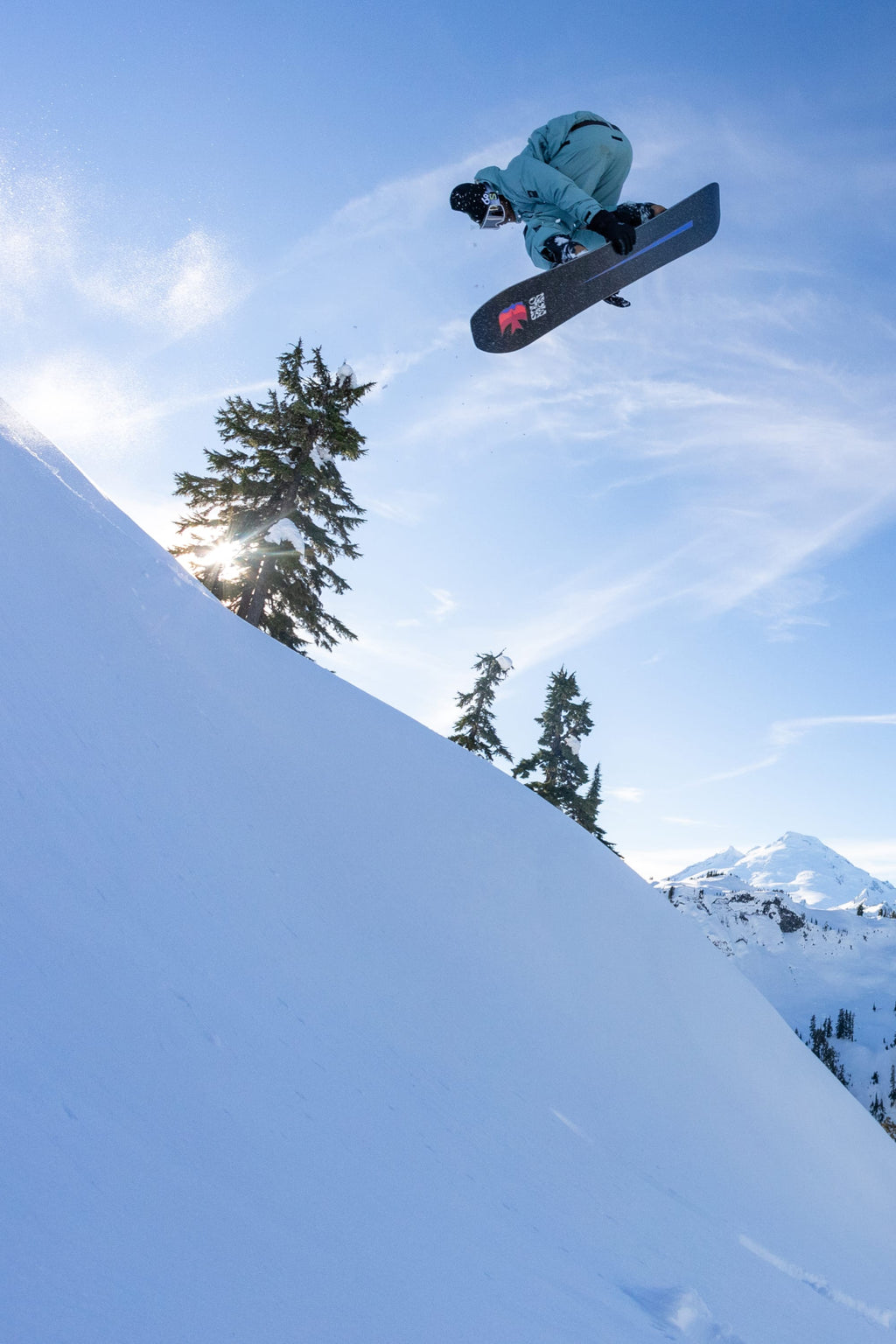 A snowboarder in light blue gear and 686 Men's GORE-TEX Dispatch™ Shell Pants performs an aerial trick off a snowy slope, framed by evergreen trees and a sunburst under a clear blue sky.