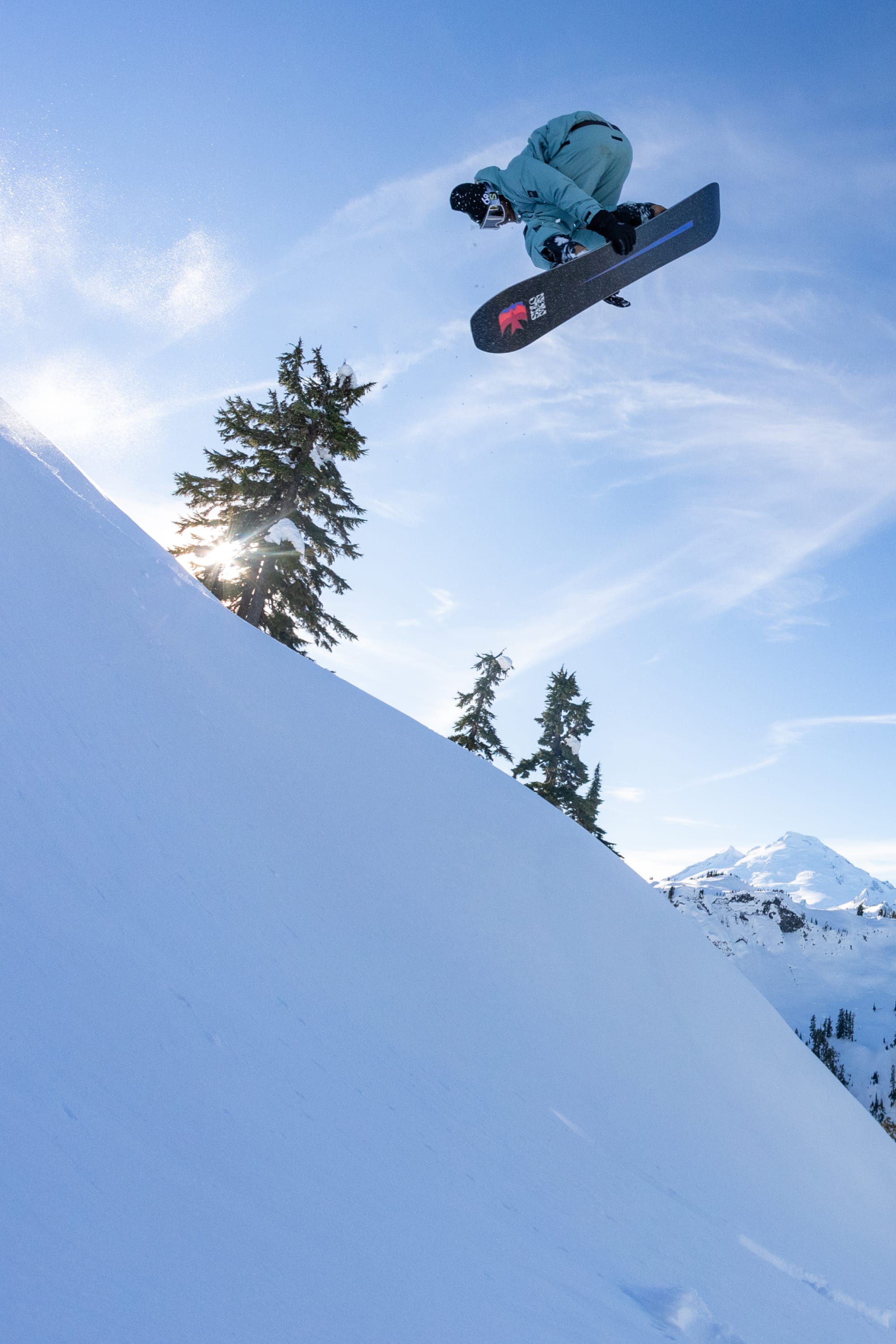 A snowboarder in light blue gear and 686 Men's GORE-TEX Dispatch™ Shell Pants performs an aerial trick off a snowy slope, framed by evergreen trees and a sunburst under a clear blue sky.