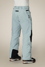 A person shown from the back wears 686 Men's GORE-TEX Dispatch™ Shell Pant in light blue with black heel patches and white-and-black boots, standing against a plain beige background.