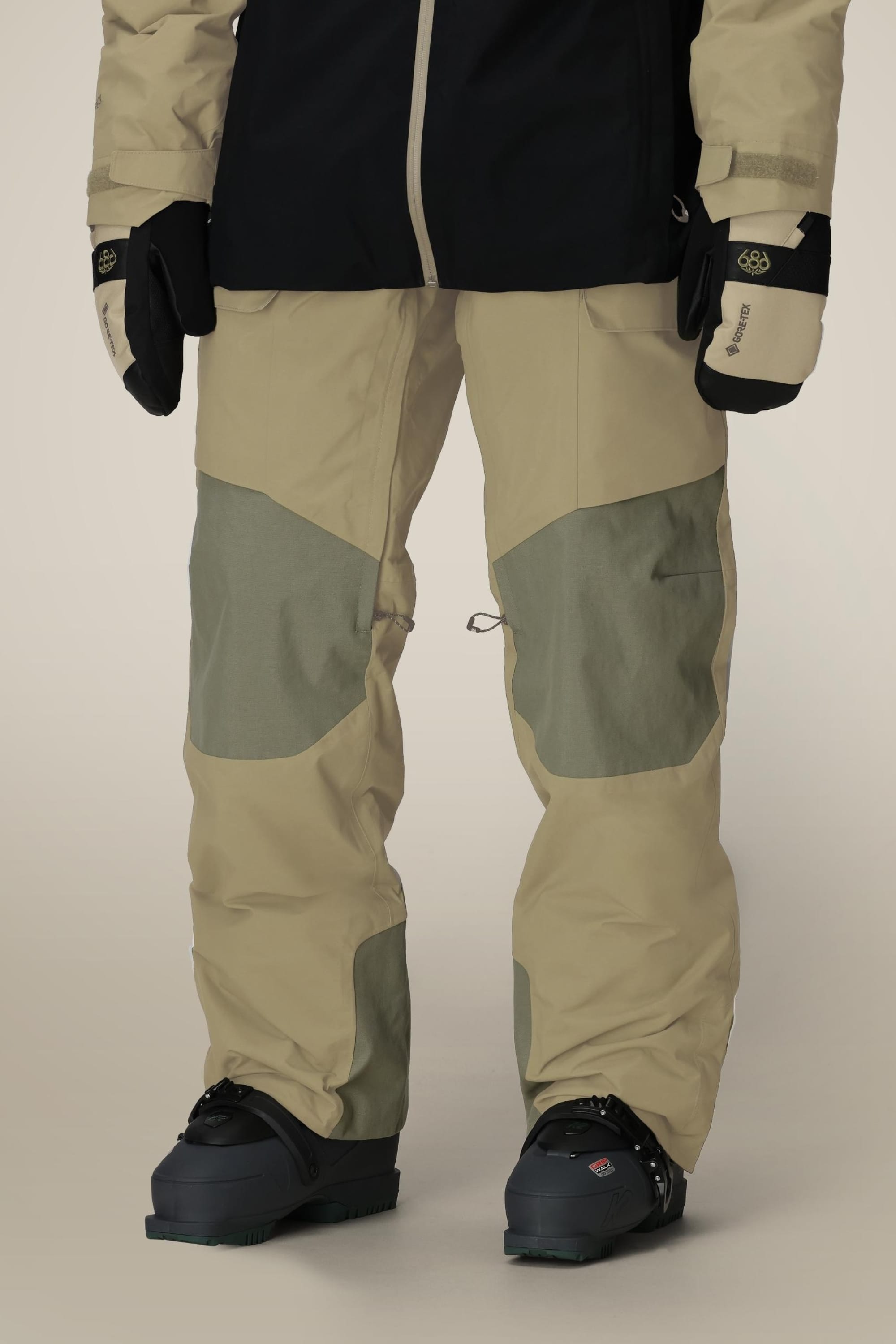 A person wearing the 686 Men's GORE-TEX Dispatch™ Shell Pant in tan and olive green with reinforced knee patches, black boots, and black-and-tan gloves, shown from shoulders down against a neutral background.