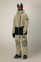 A person wearing a beige and black winter jacket, 686 Men's GORE-TEX Dispatch™ Shell Pant by 686, black gloves, dark snow boots, and ski goggles stands against a plain light background.