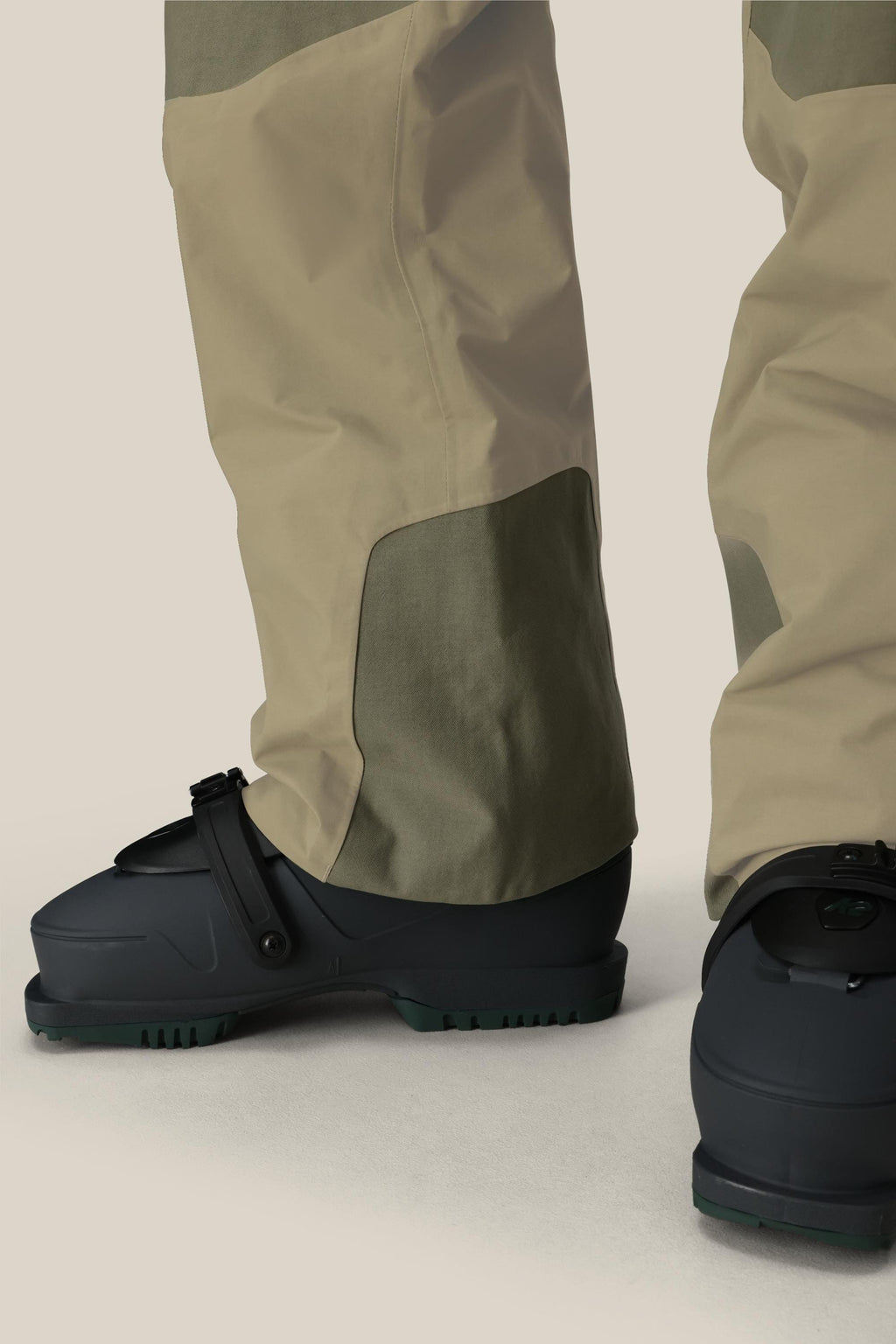 Close-up of a person wearing 686 Men's GORE-TEX Dispatch™ Shell Pant in beige and olive green, along with dark, strap-on footwear, standing on a light-gray surface. Image focuses on the lower legs and feet.
