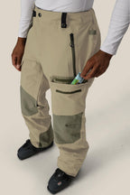 Wearing the 686 Men's GORE-TEX Dispatch™ Shell Pant in beige and olive green, along with black ski boots and a white long-sleeve shirt, a person puts a small packaged item into a zippered thigh pocket.