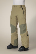 A person wears 686 Men's GORE-TEX Dispatch™ Shell Pant in beige and olive green, featuring zippered pockets and reinforced knee patches, paired with black snow boots, standing against a plain background.