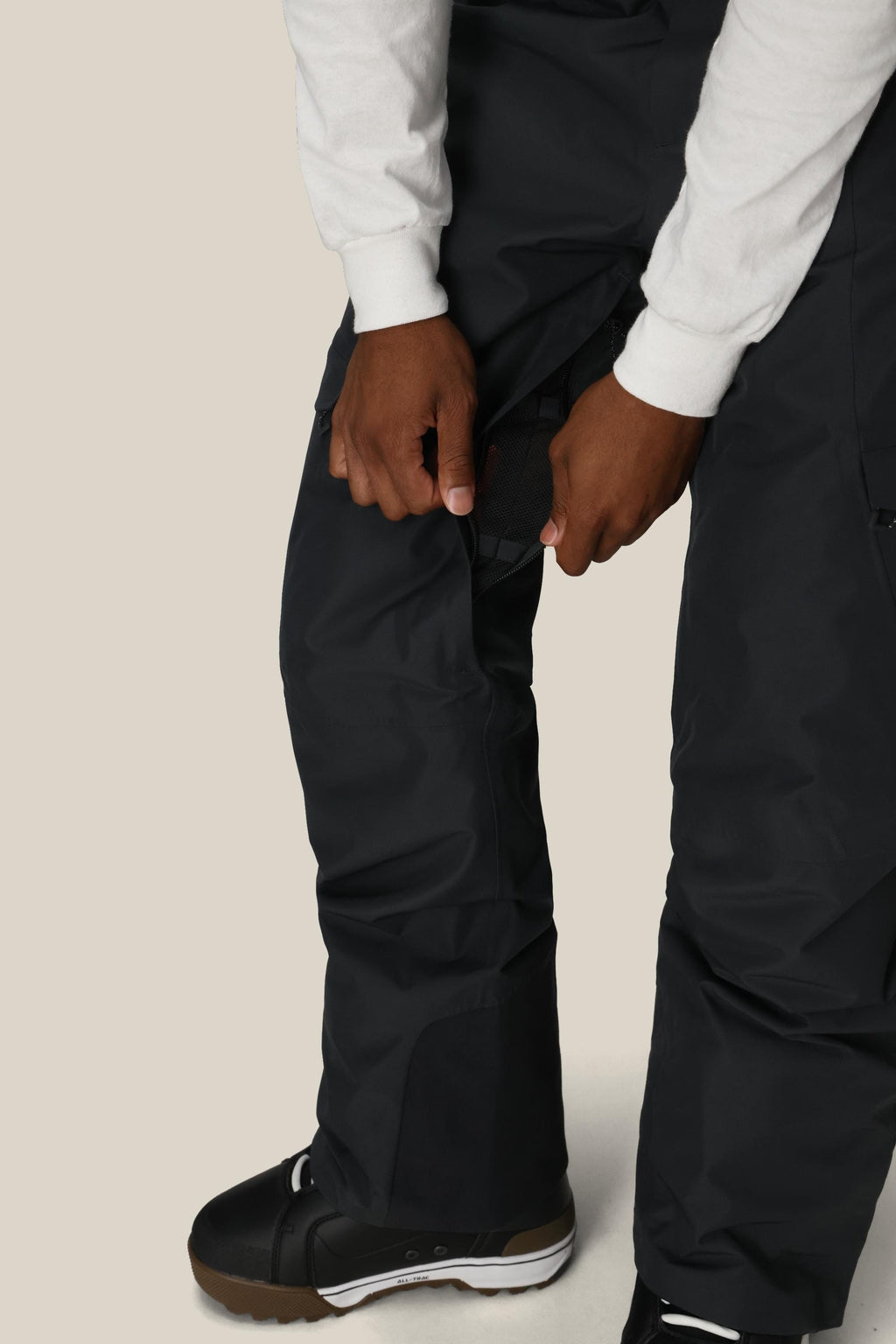 A person wearing 686 Men's Quantum Thermagraph® Pant by 686 and black winter boots adjusts a side strap on their pants. They're also wearing a white long-sleeve shirt, with the image highlighting their legs and hands.