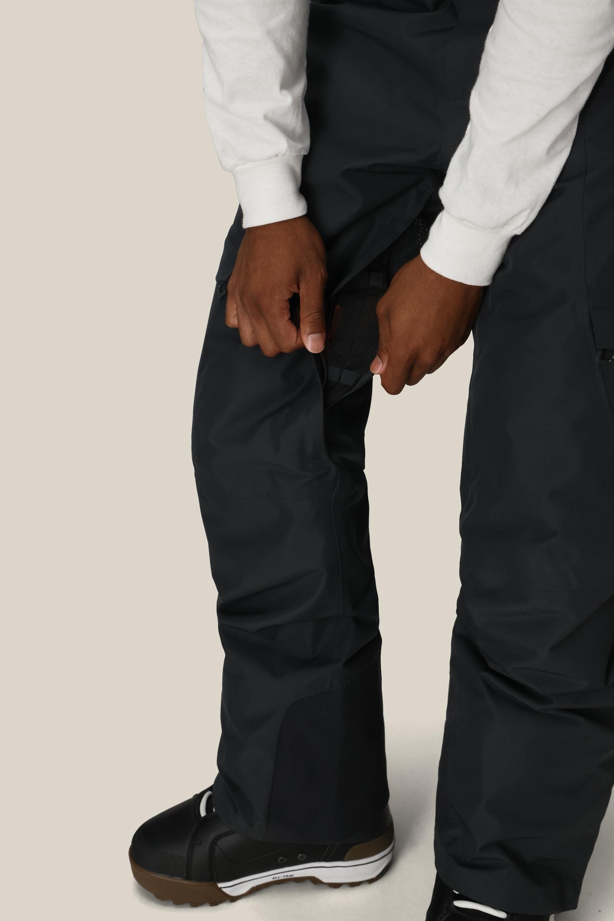 A person wearing 686 Men's Quantum Thermagraph® Pant by 686 and black winter boots adjusts a side strap on their pants. They're also wearing a white long-sleeve shirt, with the image highlighting their legs and hands.