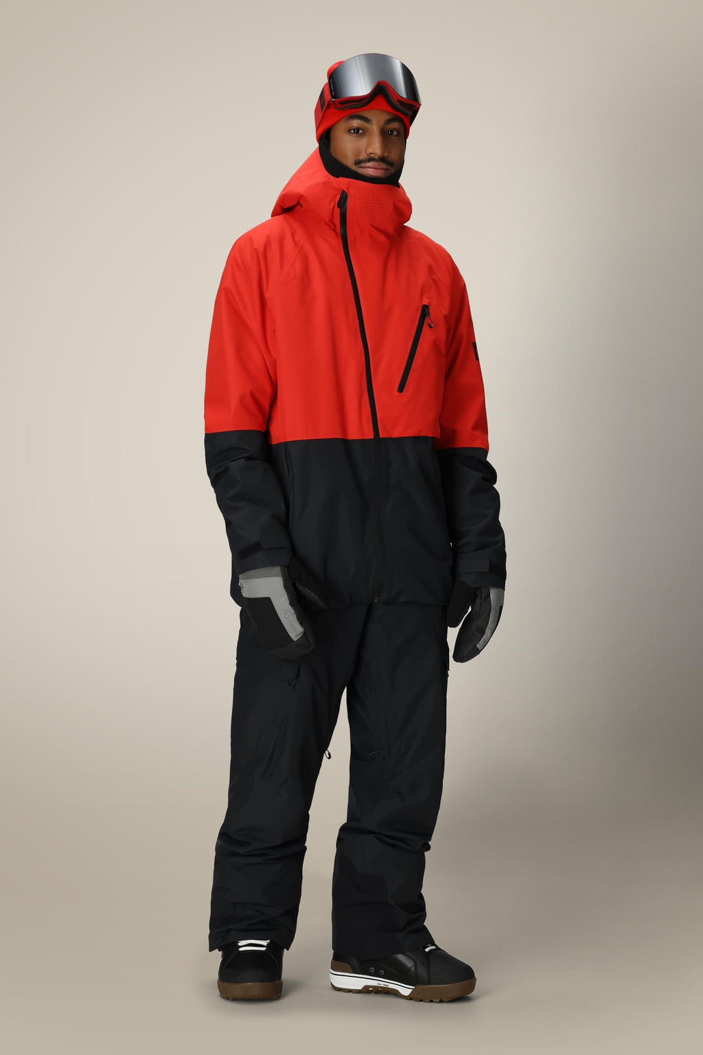 A person stands facing forward, wearing a red and black winter jacket, 686 Men's Quantum Thermagraph® Pant, gray gloves, black boots, and red ski goggles on their head against a neutral background.