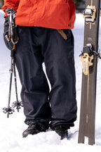A person stands in the snow wearing black 686 Dojo® GORE-TEX Pants and boots, an orange jacket, holding ski poles in one hand and a pair of skis upright in the snow with the other.