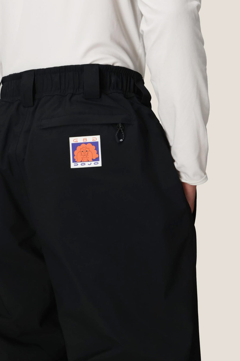 A person wears the 686 Dojo® GORE-TEX Pant by 686, featuring a colorful abstract patch on the back pocket, with a white long-sleeve shirt tucked in and their hand in the pocket.