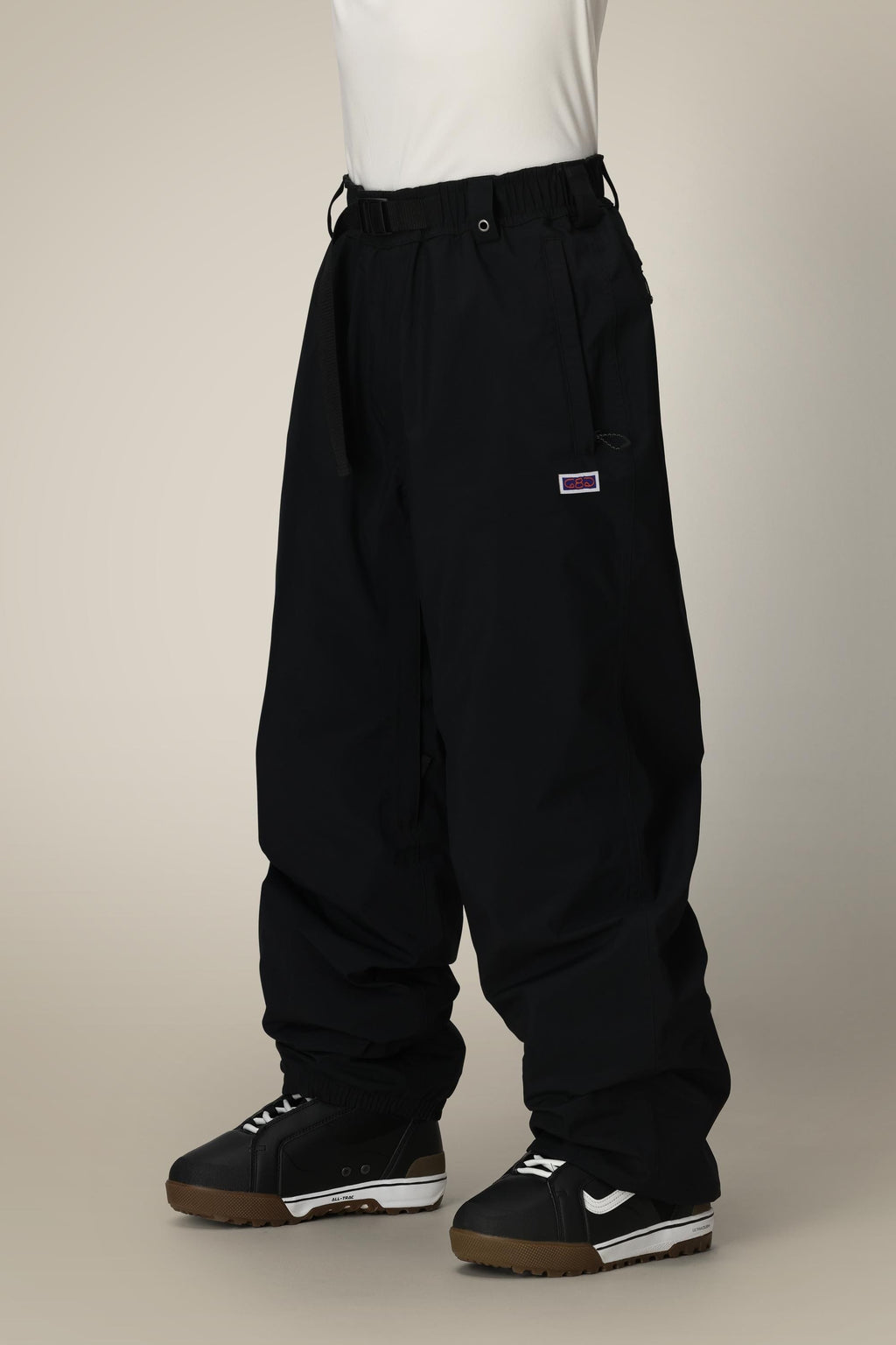 Someone stands against a plain light background wearing 686 Dojo® GORE-TEX Pant by 686—loose-fitting black nylon pants with an elastic waistband and drawstring—paired with black and white sneakers. The top half of their body is not visible.
