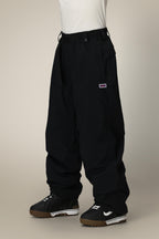 Someone stands against a plain light background wearing 686 Dojo® GORE-TEX Pant by 686—loose-fitting black nylon pants with an elastic waistband and drawstring—paired with black and white sneakers. The top half of their body is not visible.