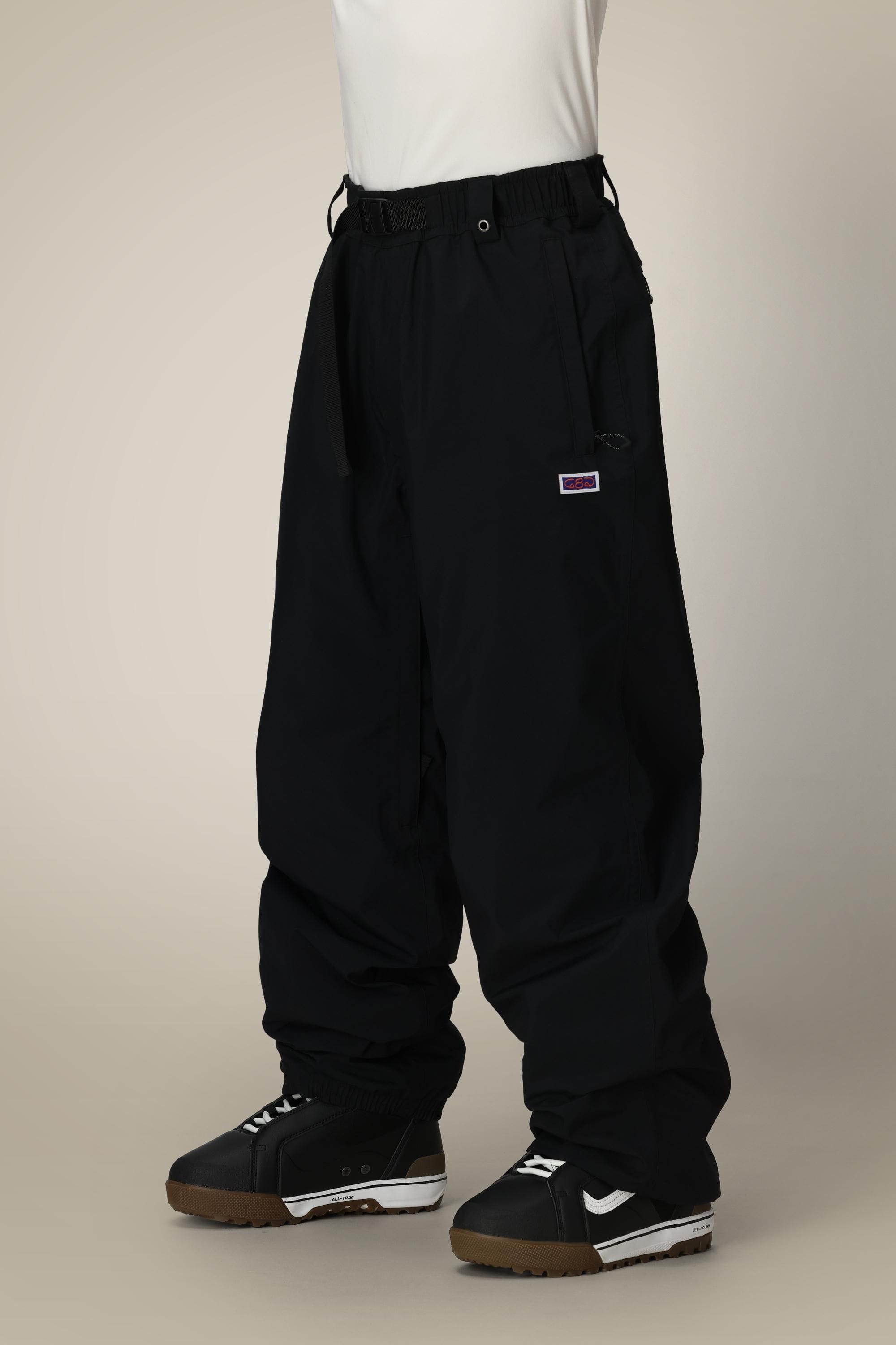 Someone stands against a plain light background wearing 686 Dojo® GORE-TEX Pant by 686—loose-fitting black nylon pants with an elastic waistband and drawstring—paired with black and white sneakers. The top half of their body is not visible.