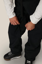 A person wearing a white long-sleeve shirt and black 686 Dojo® GORE-TEX Pants is unzipping the side of their pants. Only their torso, legs, and hands are visible, along with black and white shoes.