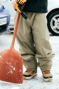 A person in tan boots, orange gloves, and 686 Dojo® GORE-TEX Pants stands on snow holding a large red snow shovel.