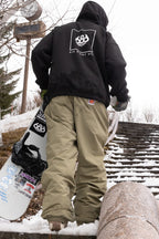 Wearing a black hoodie and 686 Dojo® GORE-TEX Pant, a person carries a snowboard up snow-dusted outdoor steps, with a clock visible on a post in the background.