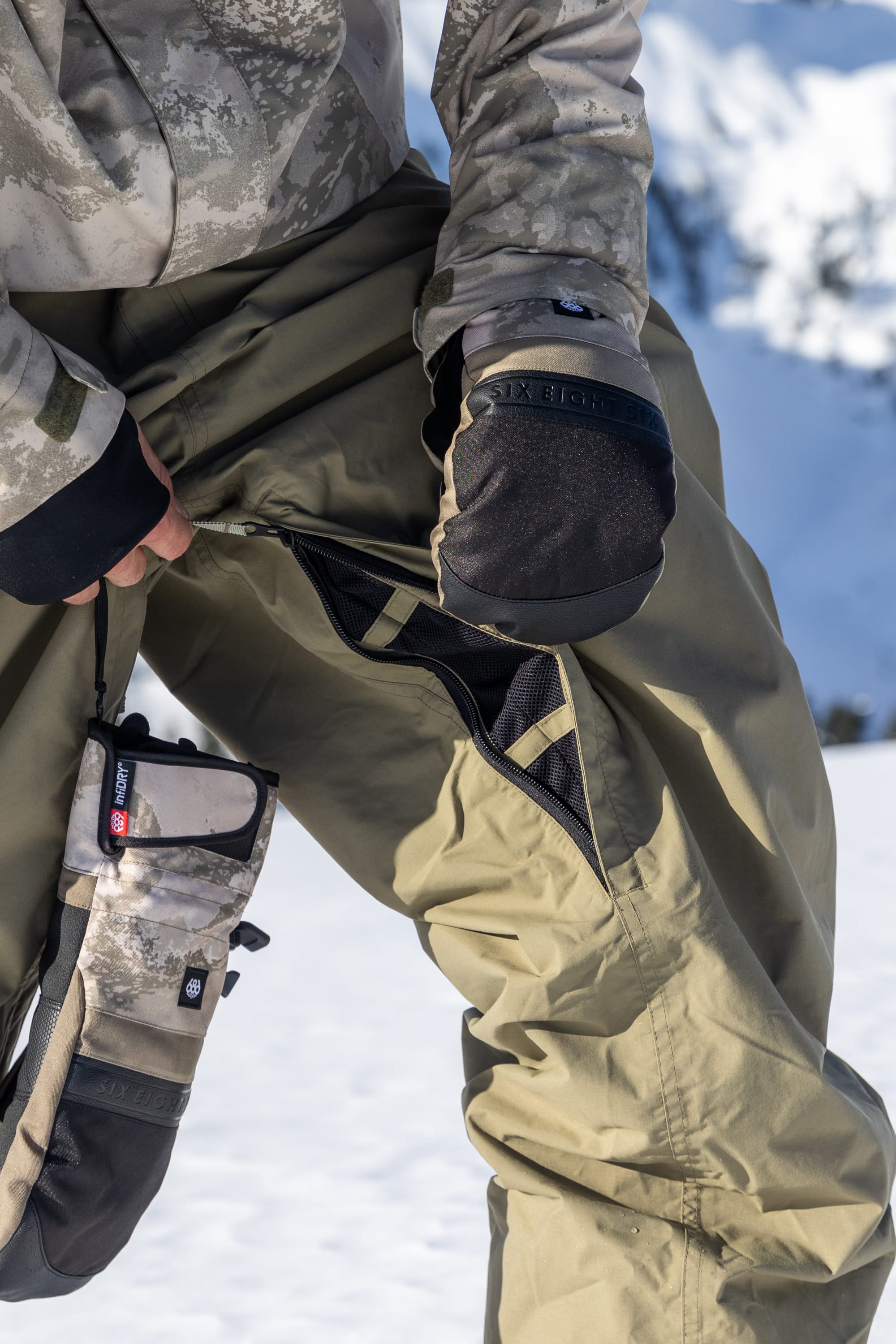 A person in camouflage winter gear unzips a vent on their olive-green 686 Dojo® GORE-TEX Pant, revealing a mesh lining. Snowy mountains and bright sunlight are visible in the background.