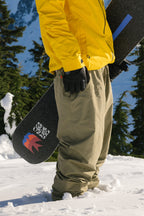 A person wearing a yellow jacket and 686 Dojo® GORE-TEX Pant by 686 stands in the snow, holding a black snowboard with colorful graphics; snowy trees and mountains are visible in the background.