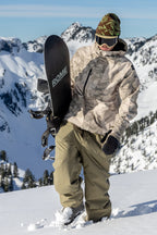 Wearing 686 Dojo® GORE-TEX Pant by 686, a person stands on a snowy mountain slope with snowboard in hand. Snow-covered peaks and a blue sky create a stunning backdrop.