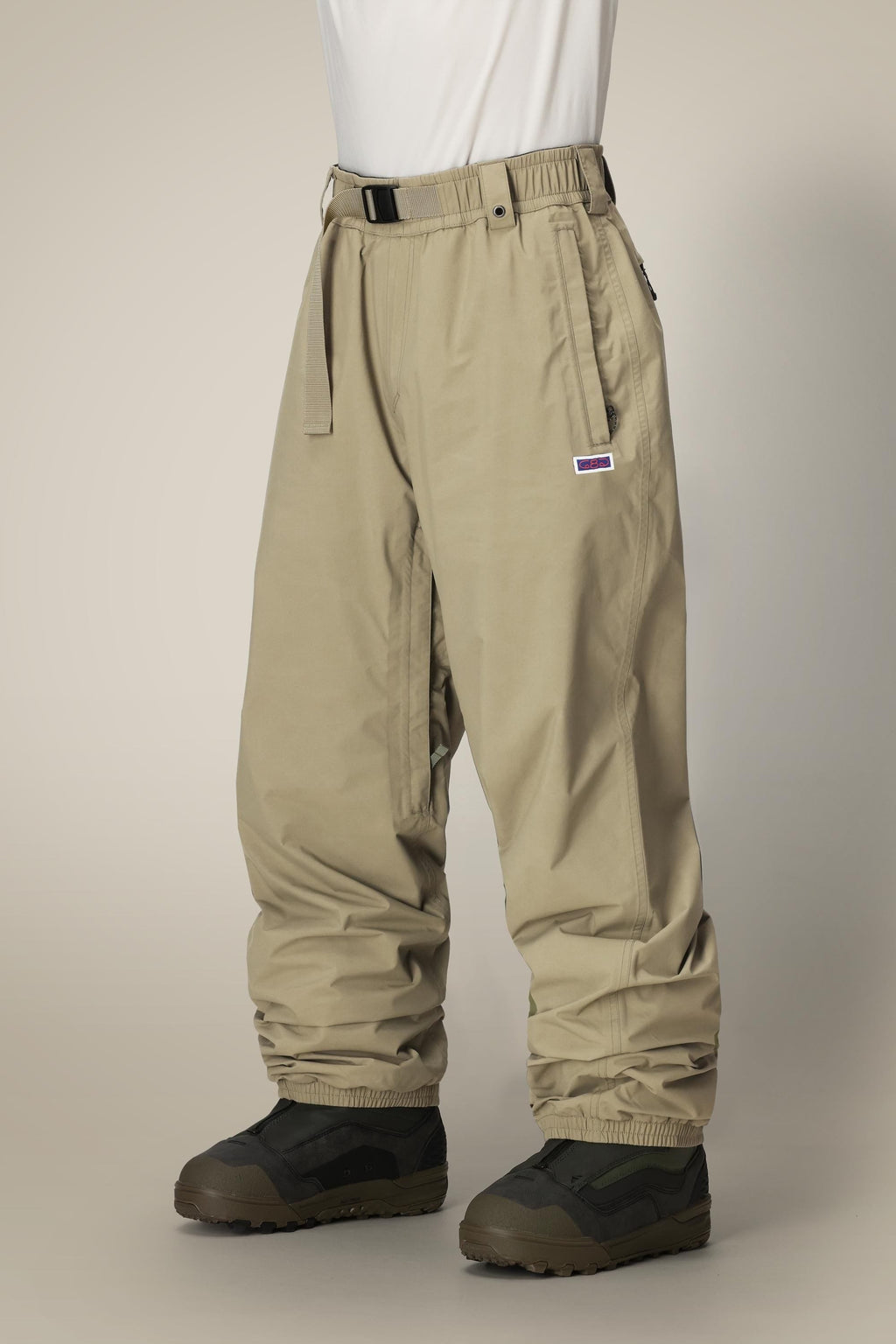 A person wearing 686 Dojo® GORE-TEX Pant by 686, tan and loose-fitting with a built-in belt, and black and olive green shoes stands against a plain light background.