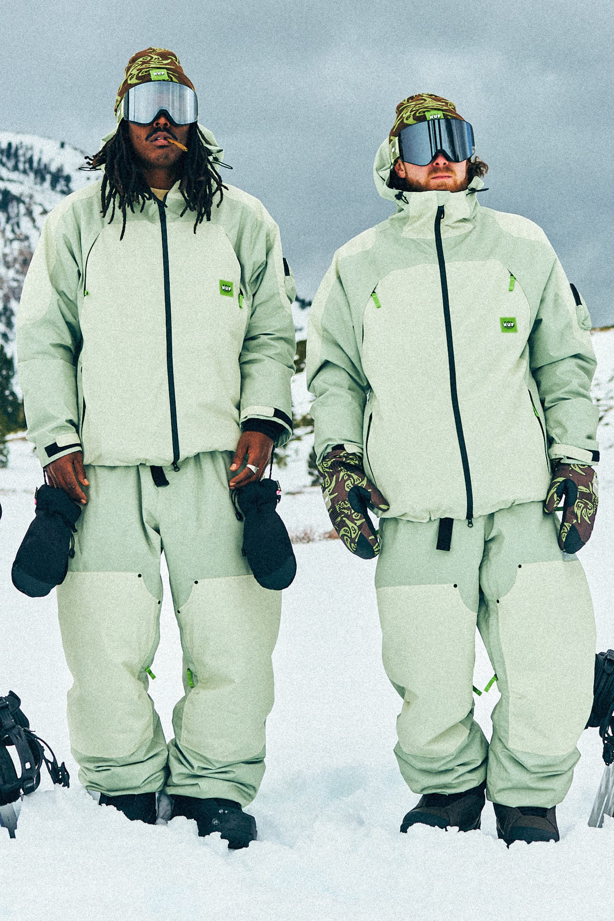 Two people stand on snow, wearing matching light jackets, gloves, goggles, and the 686 Mens HUF Double H Shell Pant. Snowy mountains and a cloudy sky create a scenic backdrop.