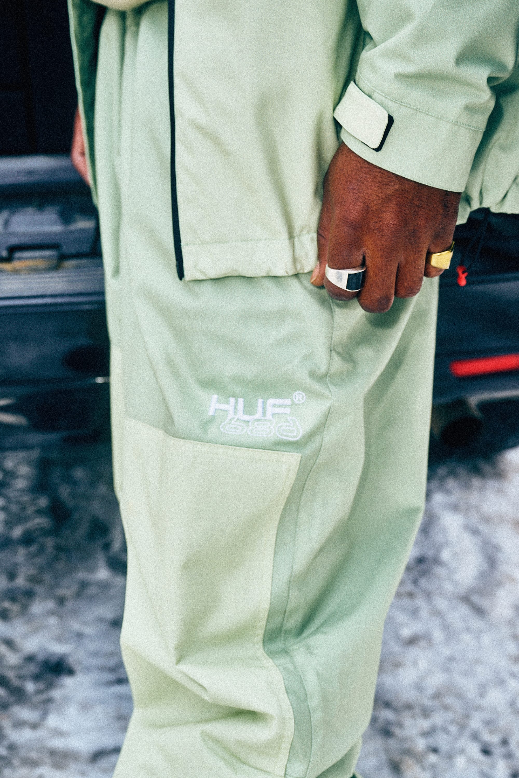 A person wearing 686 Mens HUF Double H Shell Pant in light green stands outdoors near a black vehicle with snow on the ground. Their ring-adorned hand rests by their side, and they are also wearing a matching jacket.