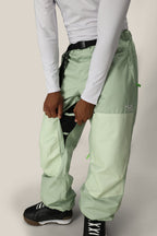 A person wearing 686 Mens HUF Double H Shell Pant in light green and a white long-sleeve shirt unzips the side to reveal a black inner layer, paired with black and white athletic shoes.