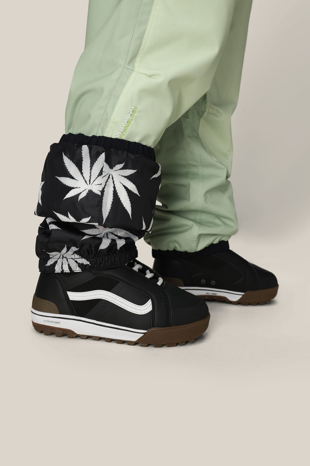 A person wearing 686 Mens HUF Double H Shell Pant in light green with black gaiters featuring white marijuana leaf prints, paired with black sneakers with white stripes and brown soles, stands against a light background.