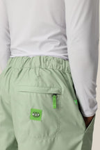A person wearing 686 Mens HUF Double H Shell Pant by 686 in light green with a back pocket and green logo patch, styled with a white long-sleeve shirt. The sporty, casual polyester look is shown cropped at the waist and lower back.