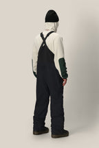 A person stands facing away, wearing a white hoodie with green patches, the 686 Men's GORE-TEX Dispatch™ Shell Bib by 686 in black, paired with black boots and a black beanie, against a plain light beige background.
