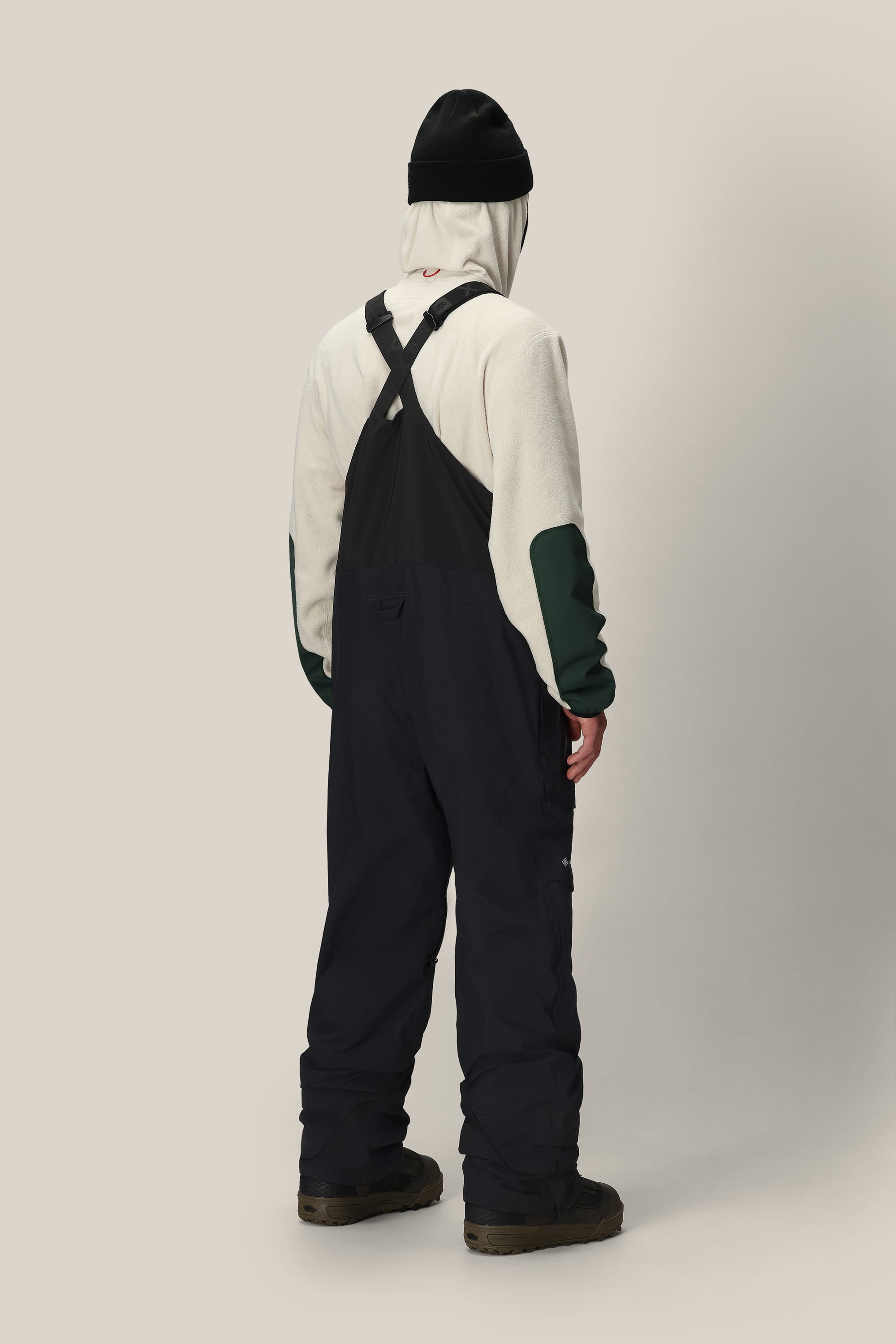 A person stands facing away, wearing a white hoodie with green patches, the 686 Men's GORE-TEX Dispatch™ Shell Bib by 686 in black, paired with black boots and a black beanie, against a plain light beige background.