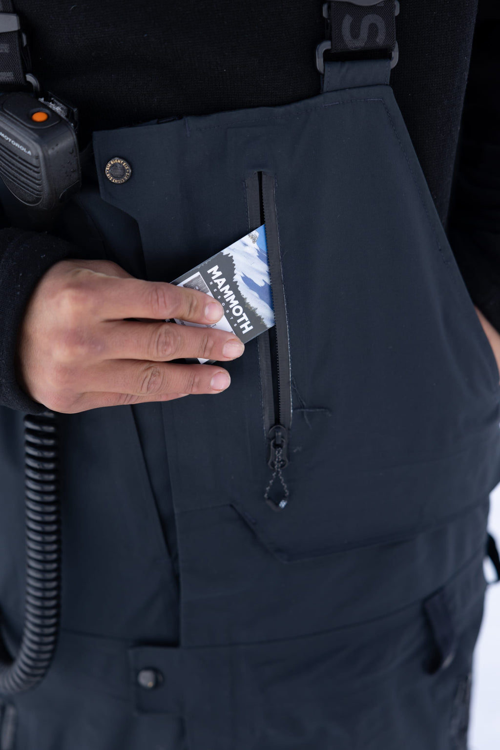 A person in a 686 Men's GORE-TEX Dispatch™ Shell Bib slips a Mammoth Mountain pass into the zippered chest pocket, with a two-way radio clipped to the shoulder.
