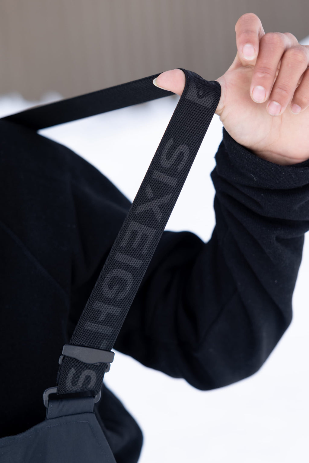 A person in a black long-sleeve shirt holds a black strap with "SIX EIGHT S" from the 686 Men's GORE-TEX Dispatch™ Shell Bib. The snowy background is blurred.